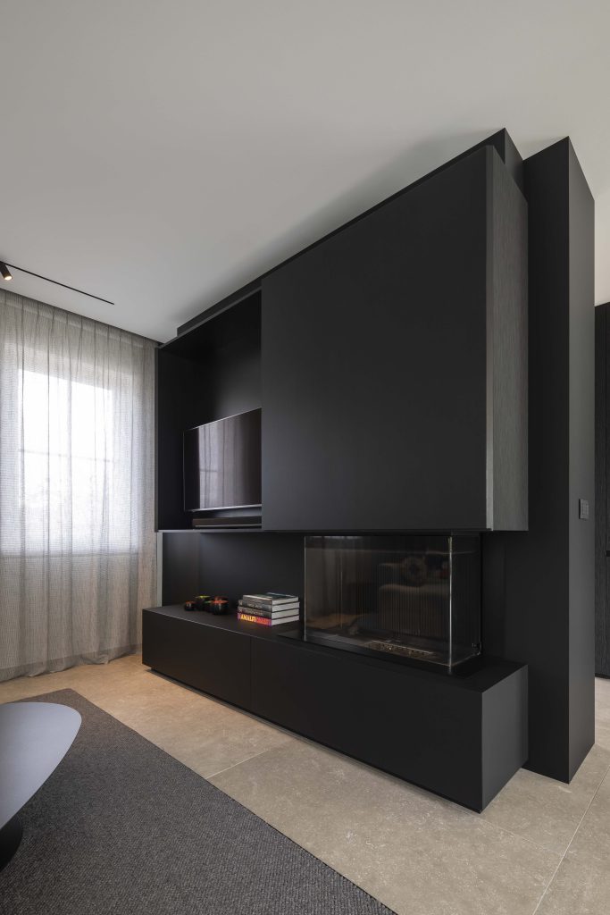 Le meuble TV est sur mesure, réalisé en noir mat à l'intérieur et en bois structuré noir pour les faces extérieures. Le meuble dévoile la télévision une fois les portes ouvertes, et la cheminée est intégrée dans le meuble